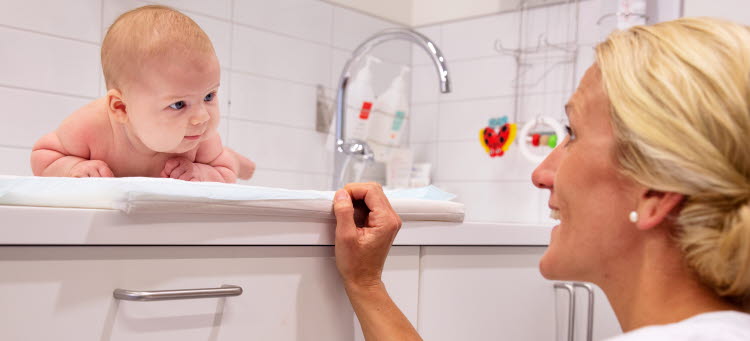 Spädbarn på en bänk som tittar på en leende kvinna.