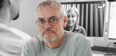 Bild på Pär Eriksson som tittar in i kameran. I bakgrunden syns en datorskärm där en kvinna är med i ett digitalt möte.