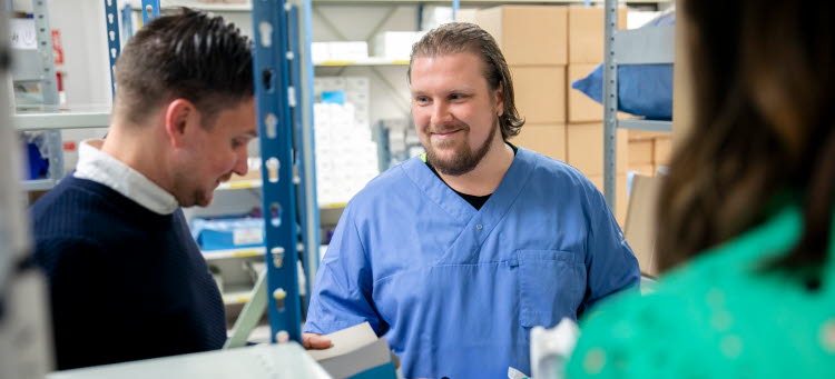 Tre personer, en vårdklädd, i ett förråd. 